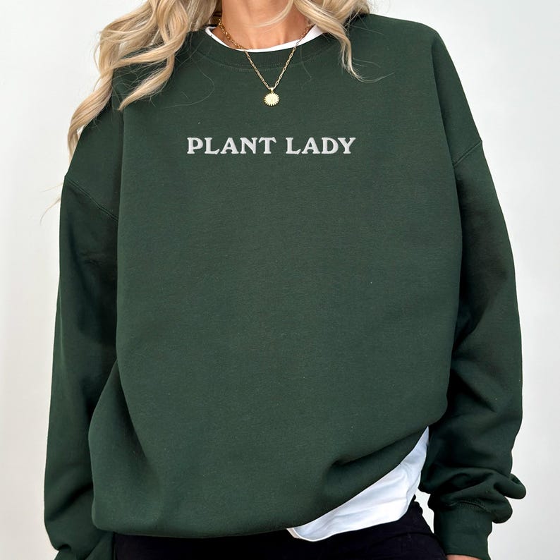 Pode incluir: Um moletom verde escuro com o texto "PLANT LADY" em letras brancas. O moletom tem gola redonda e mangas compridas. Uma camiseta branca &eacute; vis&iacute;vel por baixo.
