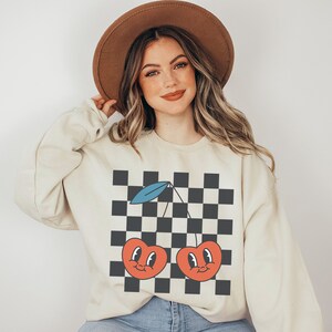 May include: A cream-colored sweatshirt with a black and white checkered pattern and a graphic of two red cherries with smiling faces. The cherries have blue stems and green leaves.