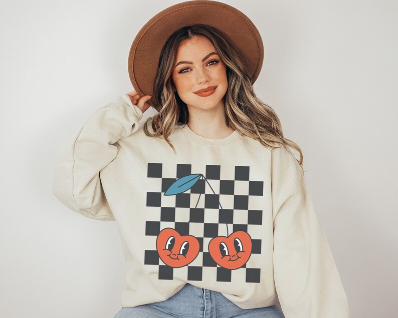 May include: A cream-colored sweatshirt with a black and white checkered pattern and a graphic of two red cherries with smiling faces. The cherries have blue stems and green leaves.