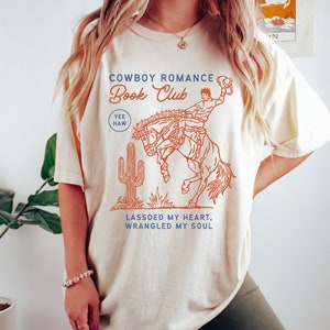 May include: A cream colored t-shirt with a graphic of a cowboy riding a bucking horse. The text "Cowboy Romance Book Club" is printed in blue and orange. The text "Yee Haw" is printed in blue. The text "Lassoed my heart, wrangled my soul" is printed in blue.