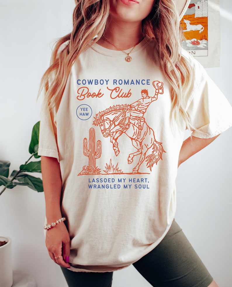 May include: A cream colored t-shirt with a graphic of a cowboy riding a bucking horse. The text "Cowboy Romance Book Club" is printed in blue and orange. The text "Yee Haw" is printed in blue. The text "Lassoed my heart, wrangled my soul" is printed in blue.
