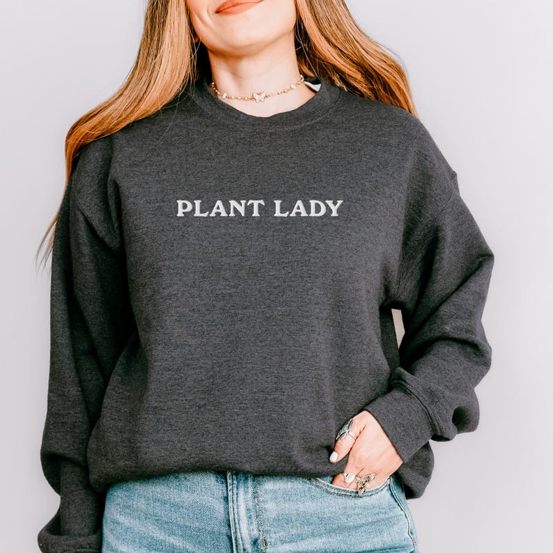 Pode incluir: Um moletom cinza escuro com o texto branco "PLANT LADY". O moletom de manga comprida tem gola redonda. A pessoa na imagem est&aacute; usando jeans azul e um colar.