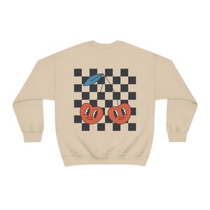 K&ouml;nnte beinhalten: Ein beiger Pullover mit einem karierten Muster und zwei roten Kirschen mit l&auml;chelnden Gesichtern auf der R&uuml;ckseite.