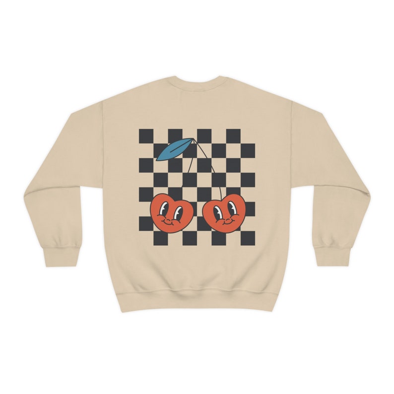 K&ouml;nnte beinhalten: Ein beiger Pullover mit einem karierten Muster und zwei roten Kirschen mit l&auml;chelnden Gesichtern auf der R&uuml;ckseite.