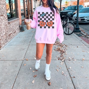 May include: A pink sweatshirt with a black and white checkered pattern and a graphic of two cherries with smiling faces.