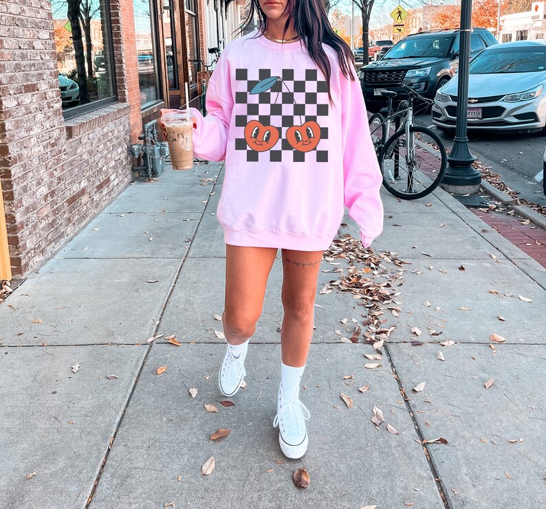 May include: A pink sweatshirt with a black and white checkered pattern and a graphic of two cherries with smiling faces.