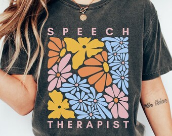 May include: A charcoal gray t-shirt with a floral design and the text "SPEECH THERAPIST" printed on the front.