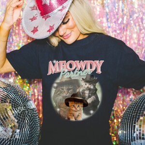 以下が含まれることがあります： 「MEOWDY Partner」とピンクのグラフィックが書かれた黒いTシャツ。カウボーイハットをかぶった猫が3匹描かれている。猫は茶トラ、白黒猫、オレンジのトラ猫である。