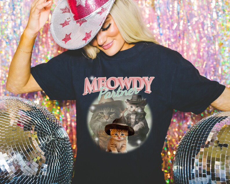 以下が含まれることがあります： 「MEOWDY Partner」とピンクのグラフィックが書かれた黒いTシャツ。カウボーイハットをかぶった猫が3匹描かれている。猫は茶トラ、白黒猫、オレンジのトラ猫である。