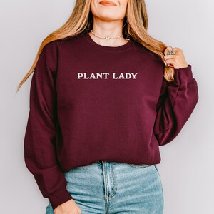 Pode incluir: Um moletom bord&ocirc; com a frase branca "PLANT LADY". O moletom &eacute; usado com jeans azuis. A imagem &eacute; um close-up da parte superior do corpo.