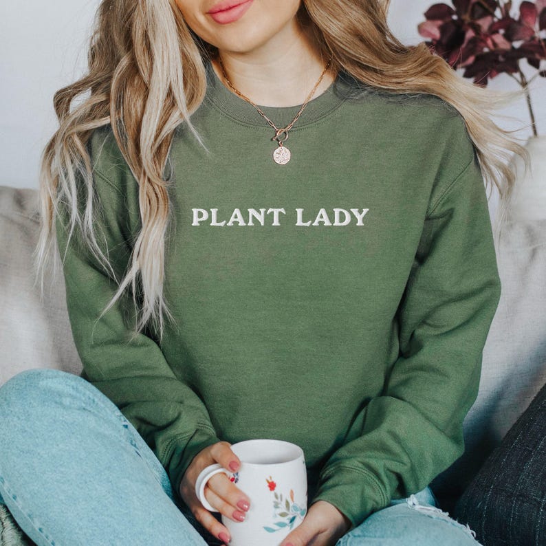 Pode incluir: Um moletom verde com o texto "PLANT LADY" em branco. A pessoa est&aacute; usando um colar de ouro e segurando uma caneca branca com desenhos florais. O moletom &eacute; de cor s&oacute;lida e tem gola redonda.