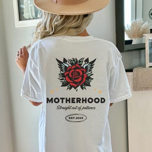 May include: White t-shirt with a red rose and wings graphic on the back. The text "MOTHERHOOD straight out of patience EST. 2025" is printed below the graphic.