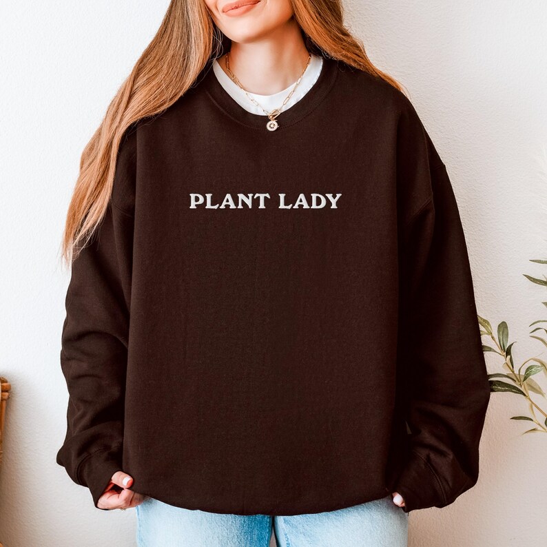 Pode incluir: Um moletom marrom escuro com o texto branco "PLANT LADY". O moletom tem gola redonda e mangas compridas. A imagem mostra uma pessoa vestindo o moletom.