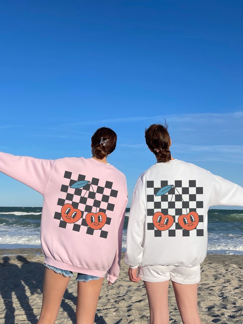 K&ouml;nnte beinhalten: Zwei Personen tragen rosa und wei&szlig;e Sweatshirts mit einem karierten Muster und einem Motiv von zwei Kirschen mit Gesichtern. Die Sweatshirts werden &uuml;ber Shorts getragen und die Personen stehen an einem Strand.