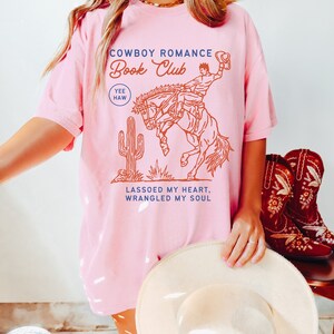 May include: Pink t-shirt with a graphic of a cowboy riding a bucking horse. The text on the shirt reads "Cowboy Romance Book Club Yee Haw Lassoed My Heart, Wrangled My Soul".