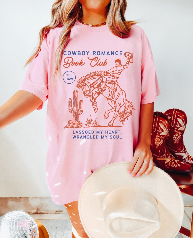 May include: Pink t-shirt with a graphic of a cowboy riding a bucking horse. The text on the shirt reads "Cowboy Romance Book Club Yee Haw Lassoed My Heart, Wrangled My Soul".
