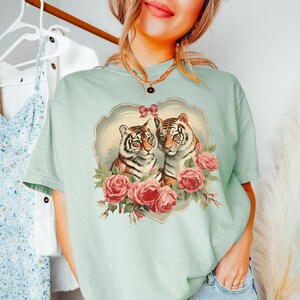 May include: A light blue t-shirt with a vintage-style illustration of two tigers with pink roses and a bow. The tigers are looking at each other and the roses are surrounding them.