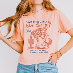 May include: Peach-colored t-shirt with a cowboy-themed graphic. The design features a cowboy riding a bucking bronco, a cactus, and the text "Cowboy Romance Book Club." Additional text reads "Lassoed My Heart, Wrangled My Soul."