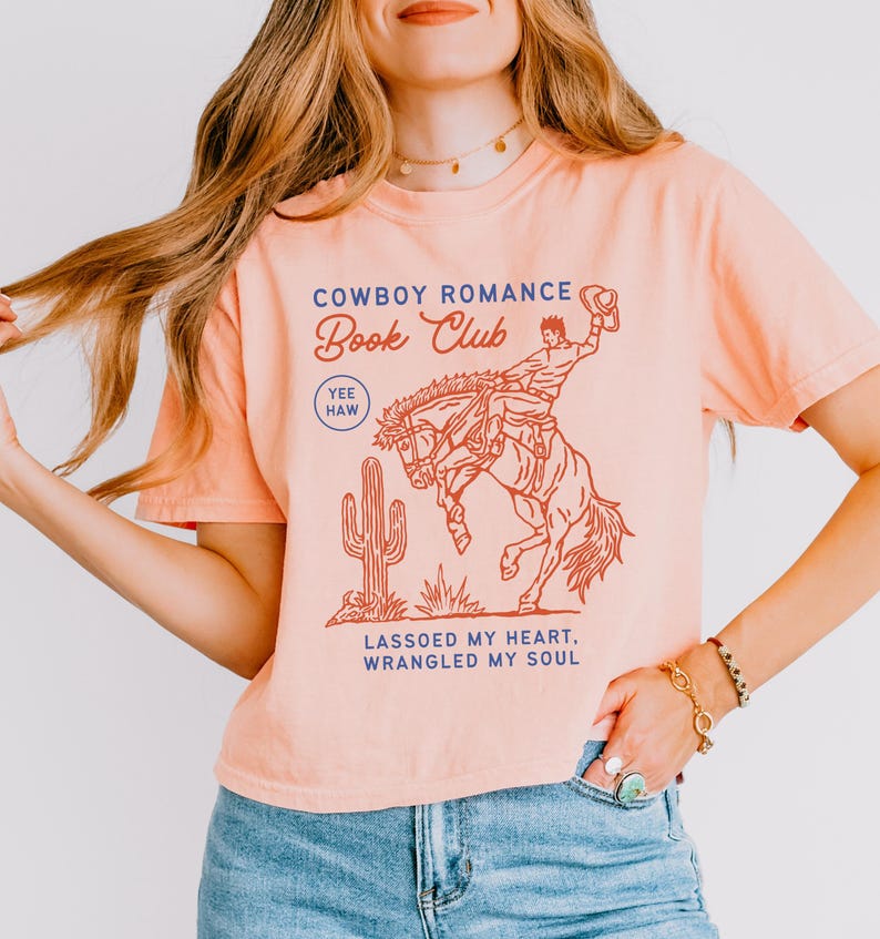 May include: Peach-colored t-shirt with a cowboy-themed graphic. The design features a cowboy riding a bucking bronco, a cactus, and the text "Cowboy Romance Book Club." Additional text reads "Lassoed My Heart, Wrangled My Soul."