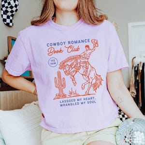 May include: A light purple t-shirt with a graphic that reads "Cowboy Romance Book Club" above an illustration of a cowboy riding a bucking bronco. The shirt also features a cactus and the phrases "Yee Haw" and "Lassoed my heart, wrangled my soul."