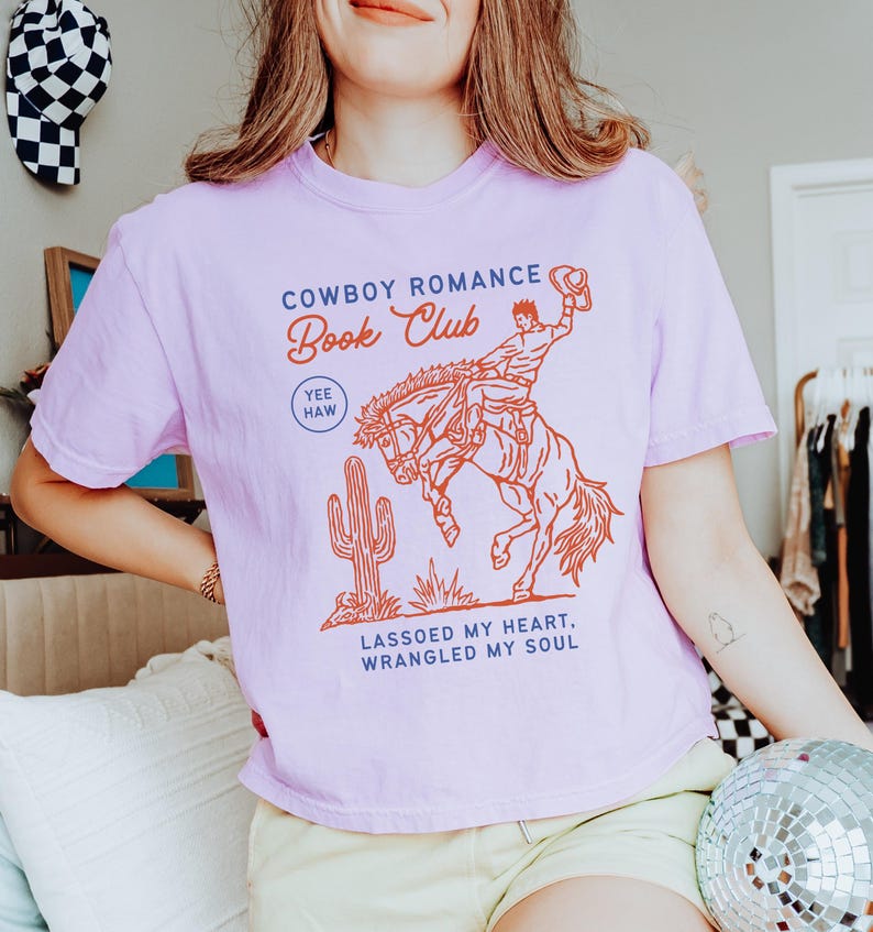 May include: A light purple t-shirt with a graphic that reads "Cowboy Romance Book Club" above an illustration of a cowboy riding a bucking bronco. The shirt also features a cactus and the phrases "Yee Haw" and "Lassoed my heart, wrangled my soul."