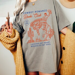 May include: Gray t-shirt with a graphic of a cowboy riding a bucking horse. The text "Cowboy Romance Book Club" is printed on the shirt, along with the words "Yee Haw" and "Lassoed my heart, wrangled my soul".