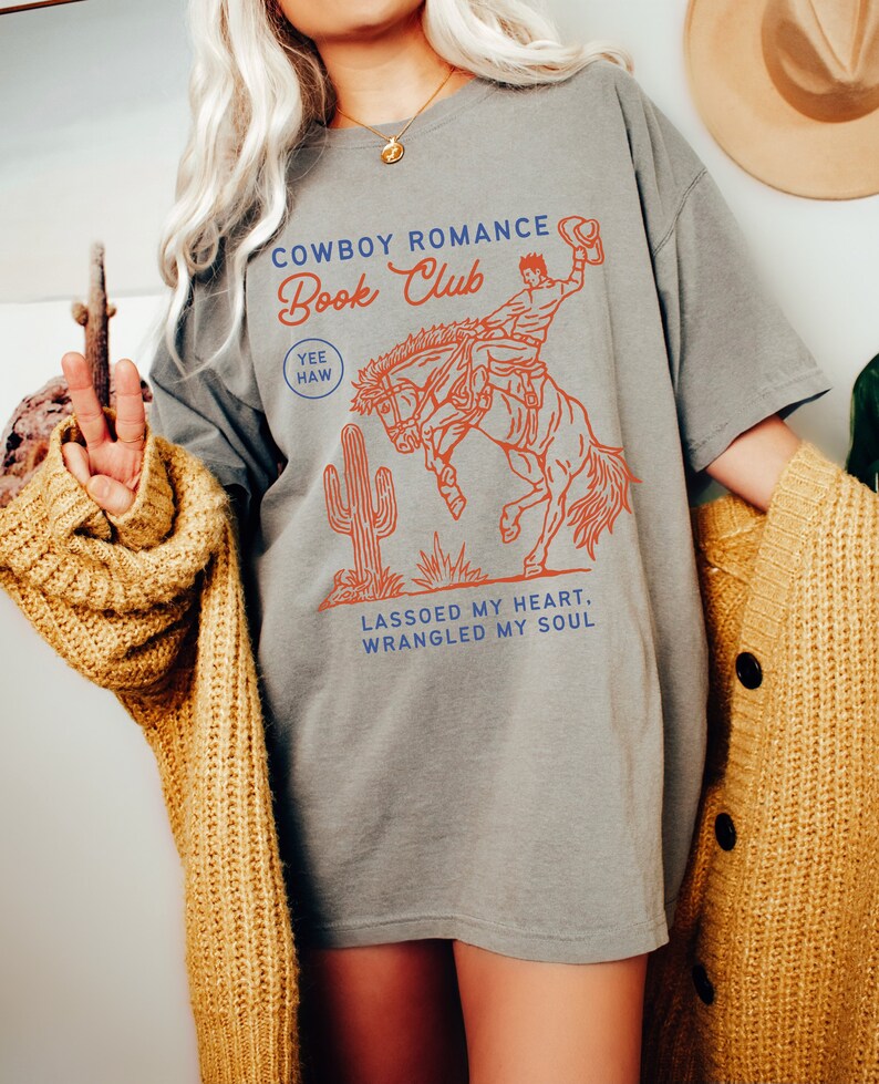 May include: Gray t-shirt with a graphic of a cowboy riding a bucking horse. The text "Cowboy Romance Book Club" is printed on the shirt, along with the words "Yee Haw" and "Lassoed my heart, wrangled my soul".