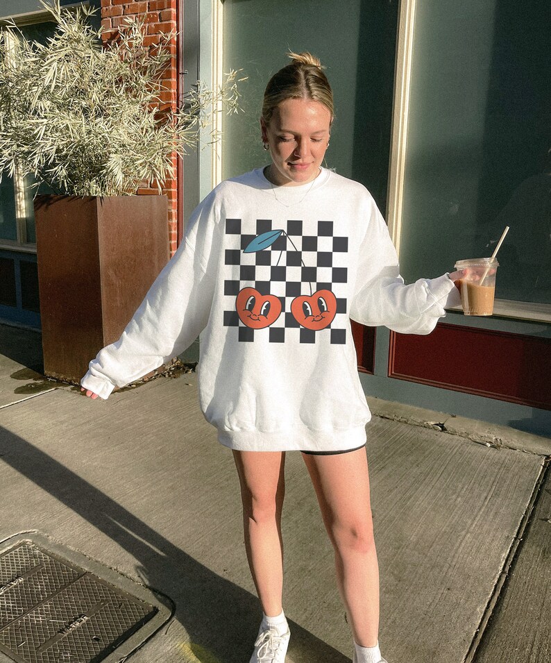 May include: White sweatshirt with a checkered pattern and a graphic of two red cherries with faces. The cherries have green stems and blue leaves.
