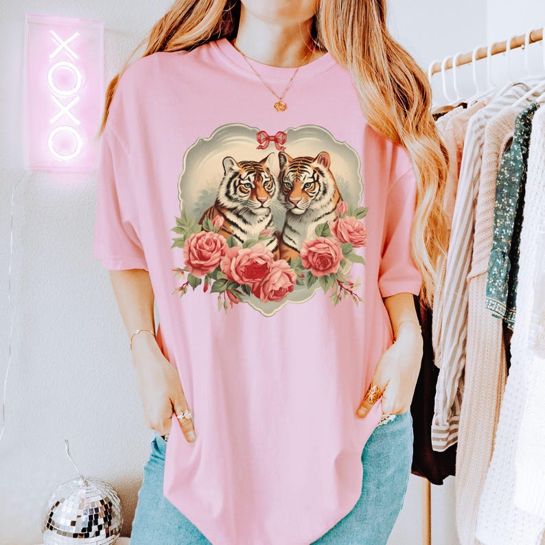 May include: A pink oversized t-shirt with a vintage-style graphic of two tigers with roses. The tigers are facing each other and have a heart-shaped design around them.