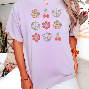 Puede incluir: Una camiseta lavanda con un dise&ntilde;o gr&aacute;fico de signos de paz, cerezas y flores en estilo retro.