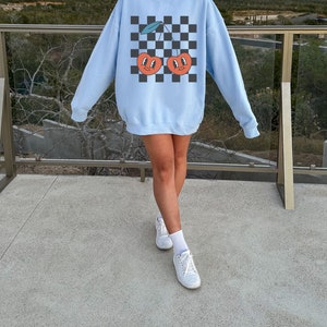 May include: A light blue sweatshirt with a checkered pattern and a graphic of two cherries with faces. The cherries are red with green stems and leaves.