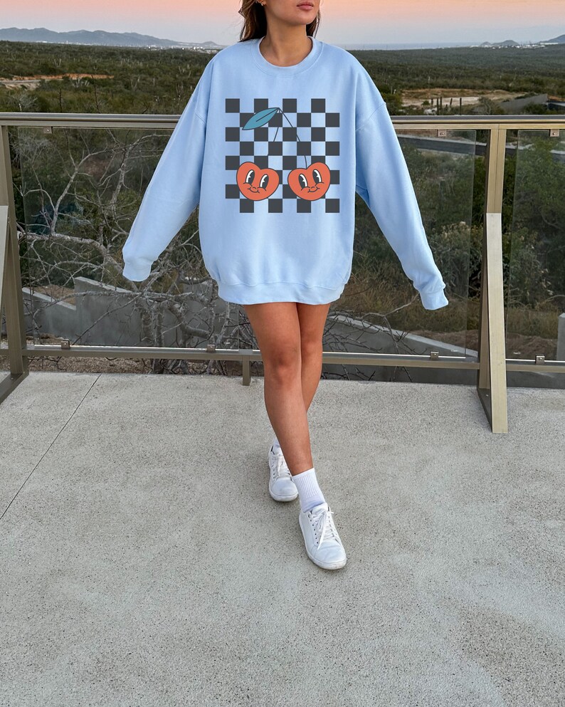 May include: A light blue sweatshirt with a checkered pattern and a graphic of two cherries with faces. The cherries are red with green stems and leaves.