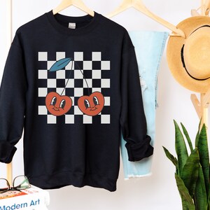 May include: Black sweatshirt with a checkered pattern and two red cherries with smiling faces. The cherries have green stems and blue leaves.
