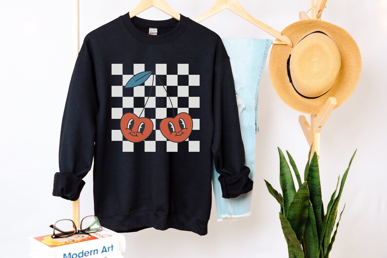 May include: Black sweatshirt with a checkered pattern and two red cherries with smiling faces. The cherries have green stems and blue leaves.