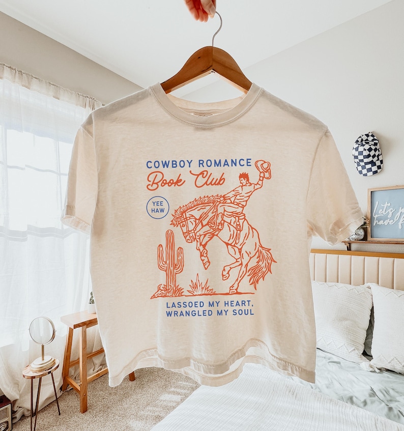 May include: Cream-colored t-shirt with a red and blue graphic. The graphic features a cowboy riding a bucking bronco, a cactus, and the text "Cowboy Romance Book Club." Additional text reads "Yee Haw" and "Lassoed My Heart, Wrangled My Soul."