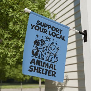 May include: A light blue flag with the text "SUPPORT YOUR LOCAL ANIMAL SHELTER" in black. The flag features a black illustration of a dog and a cat giving a high five. The flag is hanging outside.