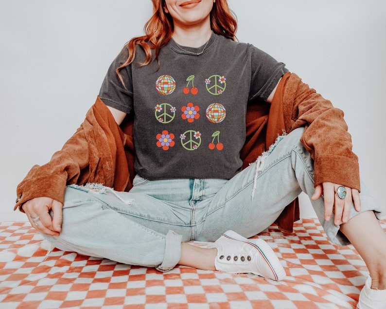 Puede incluir: Una camiseta negra con un dise&ntilde;o retro que presenta signos de paz, cerezas y bolas de discoteca. La camiseta la lleva puesta una persona sentada en una superficie a cuadros.