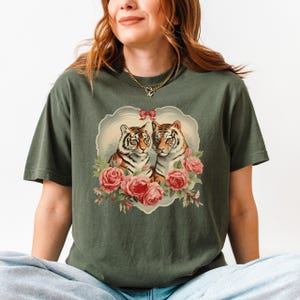 May include: A green t-shirt with a graphic of two tigers with red roses and a heart-shaped frame. The tigers are looking at each other and the roses are in bloom.