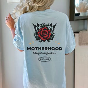 May include: Light blue t-shirt with a red rose with wings and the text "MOTHERHOOD Straight out of patience EST. 2025"