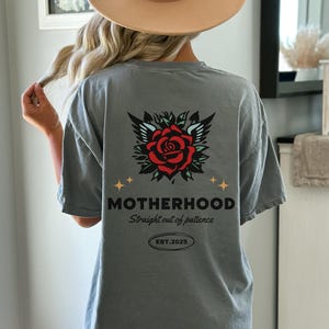 May include: A gray t-shirt with a rose and wings graphic on the back. The text "MOTHERHOOD Straight out of patience EST. 2025" is printed below the graphic.