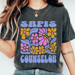 May include: Dark gray t-shirt with a floral design and the words "SAPIS COUNSELOR" in a retro font. The flowers are pink, yellow, and purple. The shirt has a vintage, worn look.