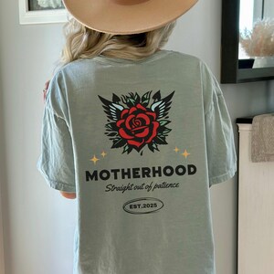 May include: A light green t-shirt with a red rose and wings graphic on the back. The text "MOTHERHOOD Straight out of patience EST. 2025" is printed below the graphic.