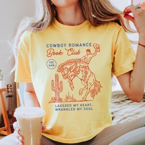 May include: Yellow t-shirt with a red and orange graphic of a cowboy riding a bucking bronco. The text on the shirt reads "Cowboy Romance Book Club" with "Yee Haw" and "Lassoed My Heart, Wrangled My Soul" also printed on the shirt.