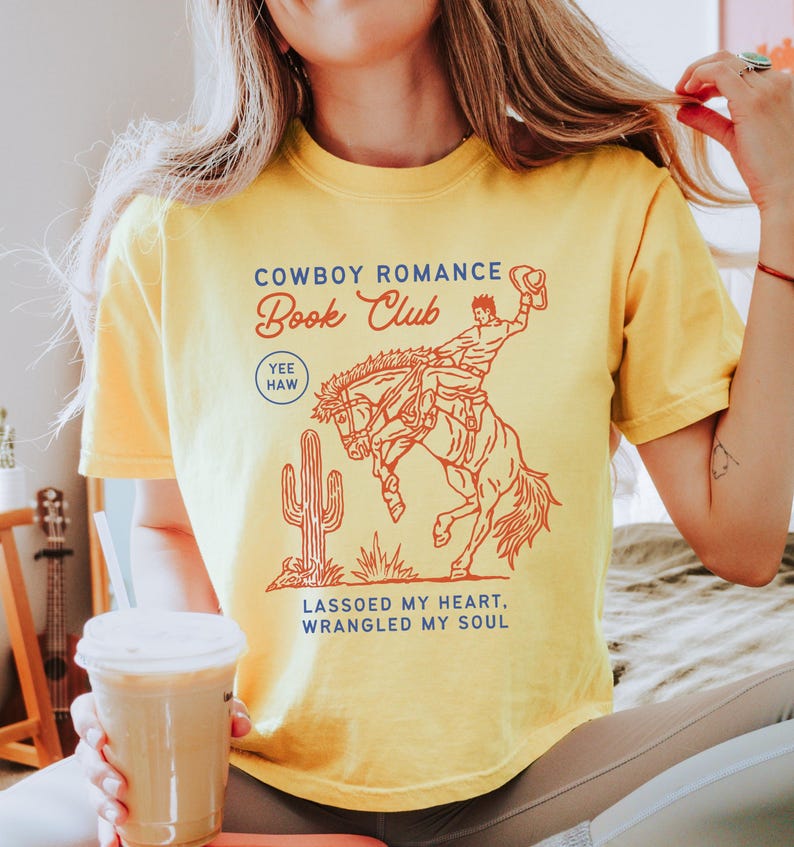 May include: Yellow t-shirt with a red and orange graphic of a cowboy riding a bucking bronco. The text on the shirt reads "Cowboy Romance Book Club" with "Yee Haw" and "Lassoed My Heart, Wrangled My Soul" also printed on the shirt.