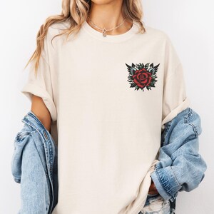 May include: A cream colored t-shirt with a red rose tattoo design on the left chest.