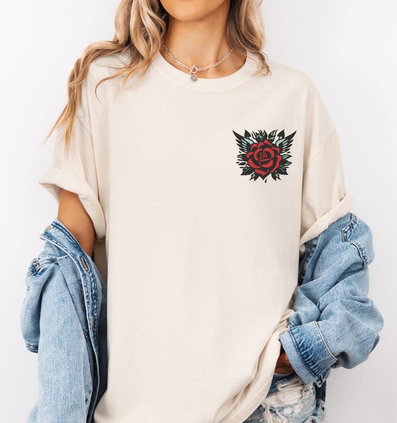 May include: A cream colored t-shirt with a red rose tattoo design on the left chest.