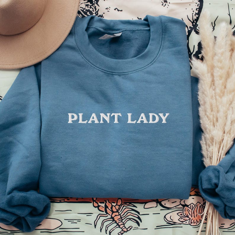 Pode incluir: Um moletom azul com o texto "PLANT LADY" em branco. Um chap&eacute;u bege e grama ornamental seca est&atilde;o no fundo. O moletom est&aacute; dobrado sobre uma superf&iacute;cie estampada.