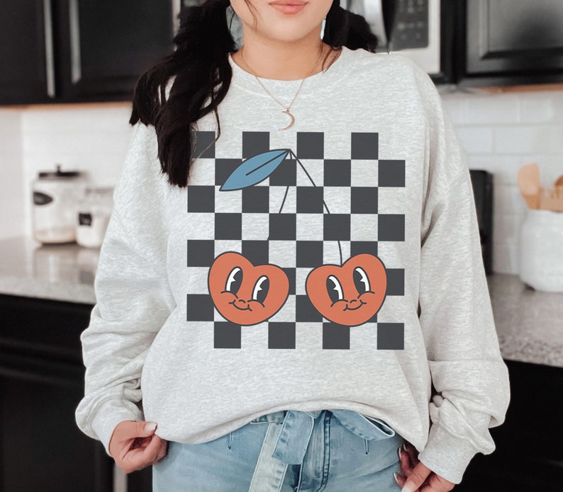 May include: A gray sweatshirt with a black and white checkered pattern and two red cherries with faces on it.