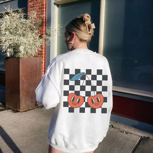 K&ouml;nnte beinhalten: Wei&szlig;er Pullover mit einem schwarz-wei&szlig;en Karomuster und einer Grafik von zwei roten Kirschen mit l&auml;chelnden Gesichtern auf der R&uuml;ckseite.