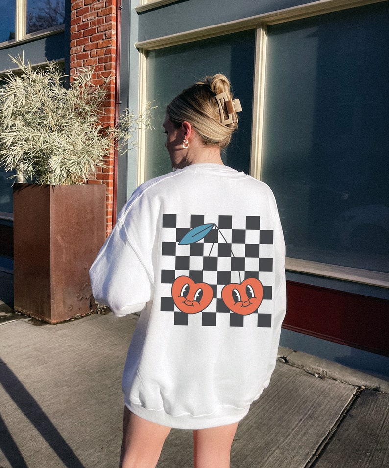 K&ouml;nnte beinhalten: Wei&szlig;er Pullover mit einem schwarz-wei&szlig;en Karomuster und einer Grafik von zwei roten Kirschen mit l&auml;chelnden Gesichtern auf der R&uuml;ckseite.
